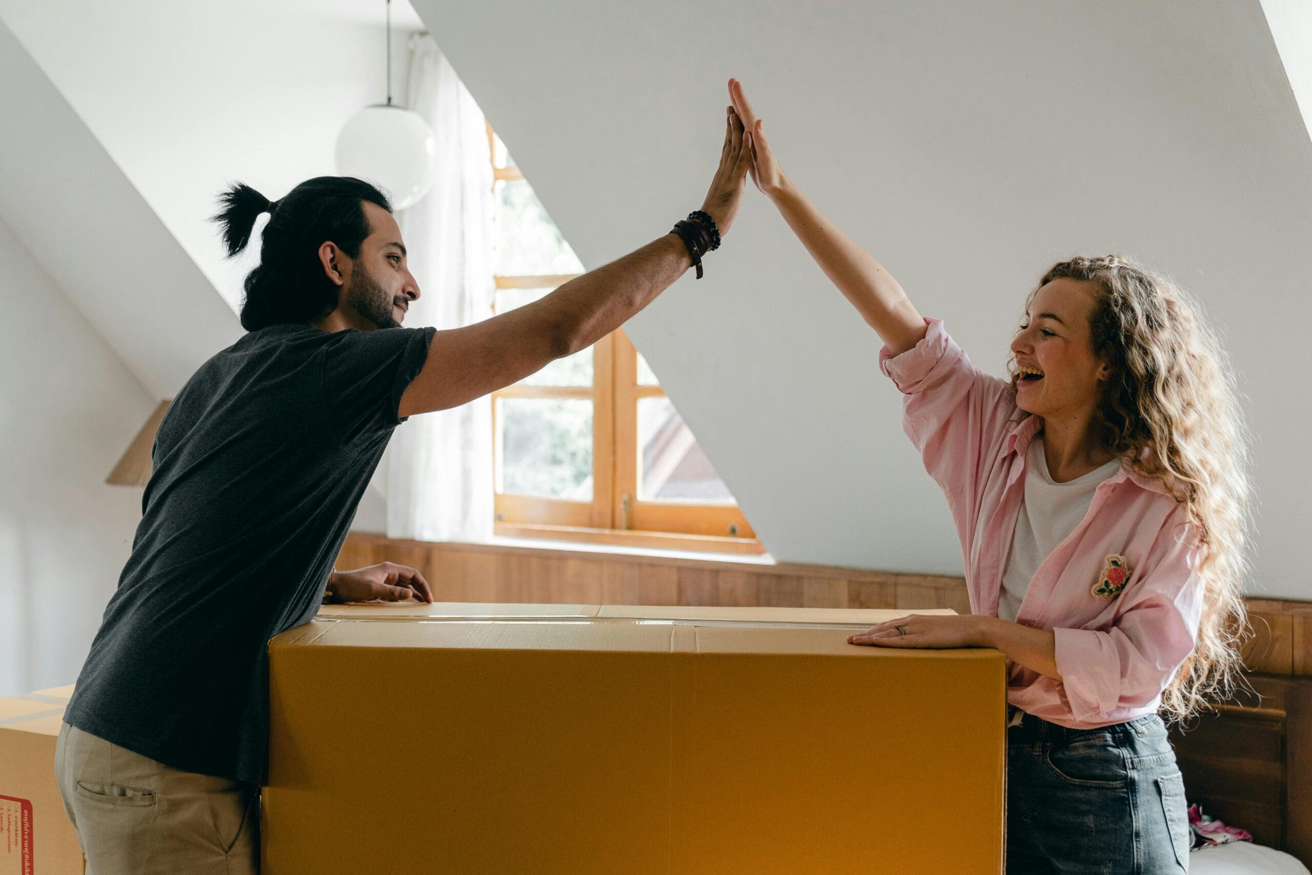 A man and a woman share a high-five over a large cardboard box in a bright room with a slanted ceiling and a window showcasing the beautiful exterior renovations of their Madison, WI home. The man has a bun hairstyle and wears a dark shirt, while the woman has curly hair and wears a pink shirt.